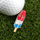 A golf ball sits on green grass next to a golf ball marker and Logie Joe's Golf Red White Blue Popsicle enamel pin.