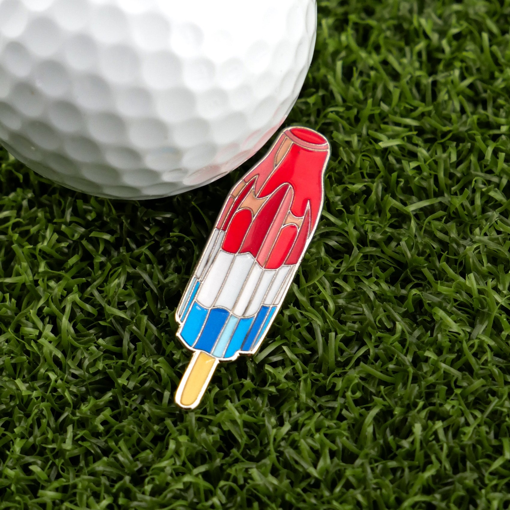 A golf ball sits on green grass next to a golf ball marker and Logie Joe's Golf Red White Blue Popsicle enamel pin.