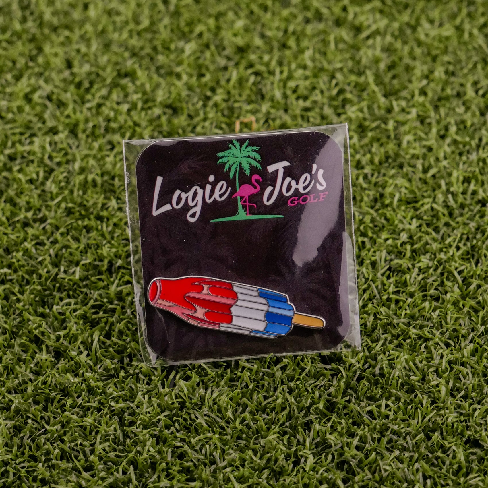The Logie Joe's Golf Red White Blue Popsicle pin, shaped like a rocket popsicle, is perfect as a golf ball marker or accessory. It comes displayed on artificial grass and attached to a black card with the Logie Joe's Golf logo.