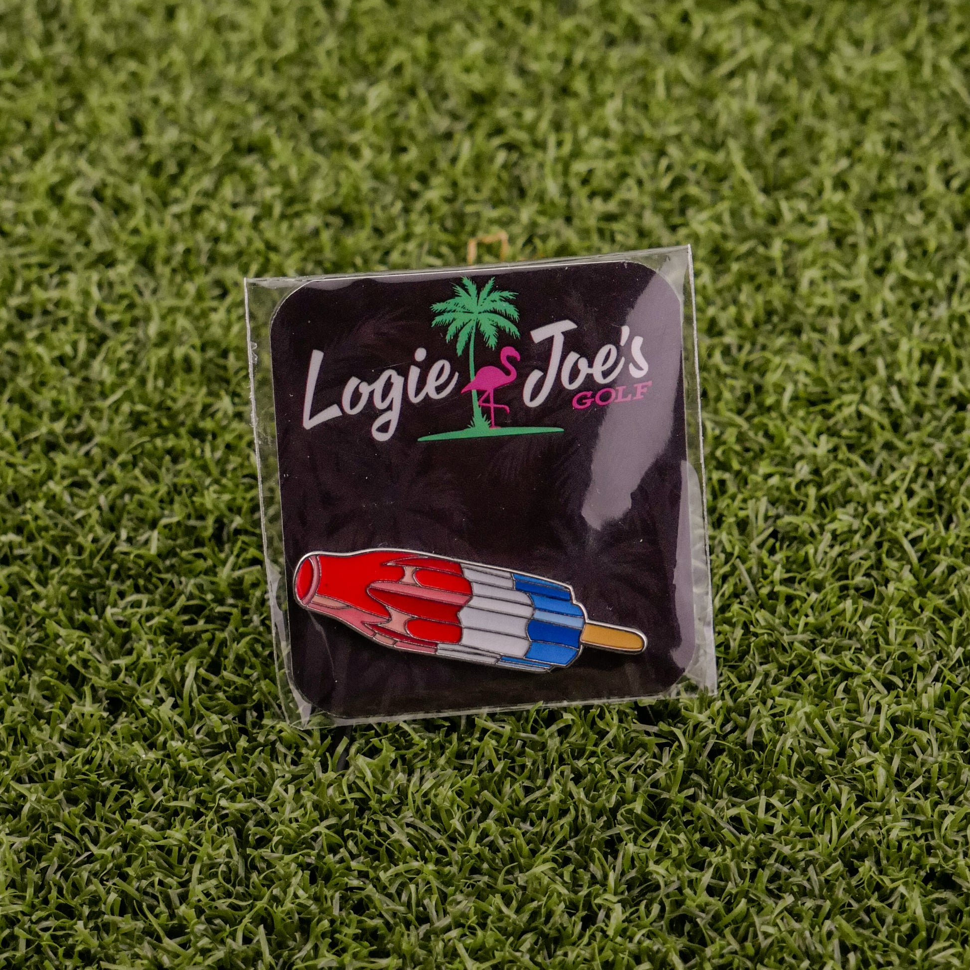 The Logie Joe's Golf Red White Blue Popsicle pin, shaped like a rocket popsicle, is perfect as a golf ball marker or accessory. It comes displayed on artificial grass and attached to a black card with the Logie Joe's Golf logo.