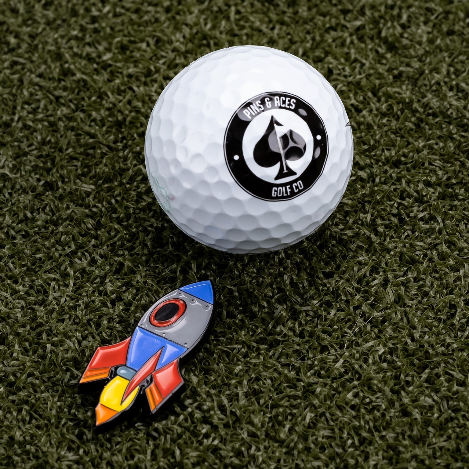 A Pins and Aces golf ball rests on green turf beside the Rocket Emoji Ball Marker, a colorful enamel marker shaped like a rocket.