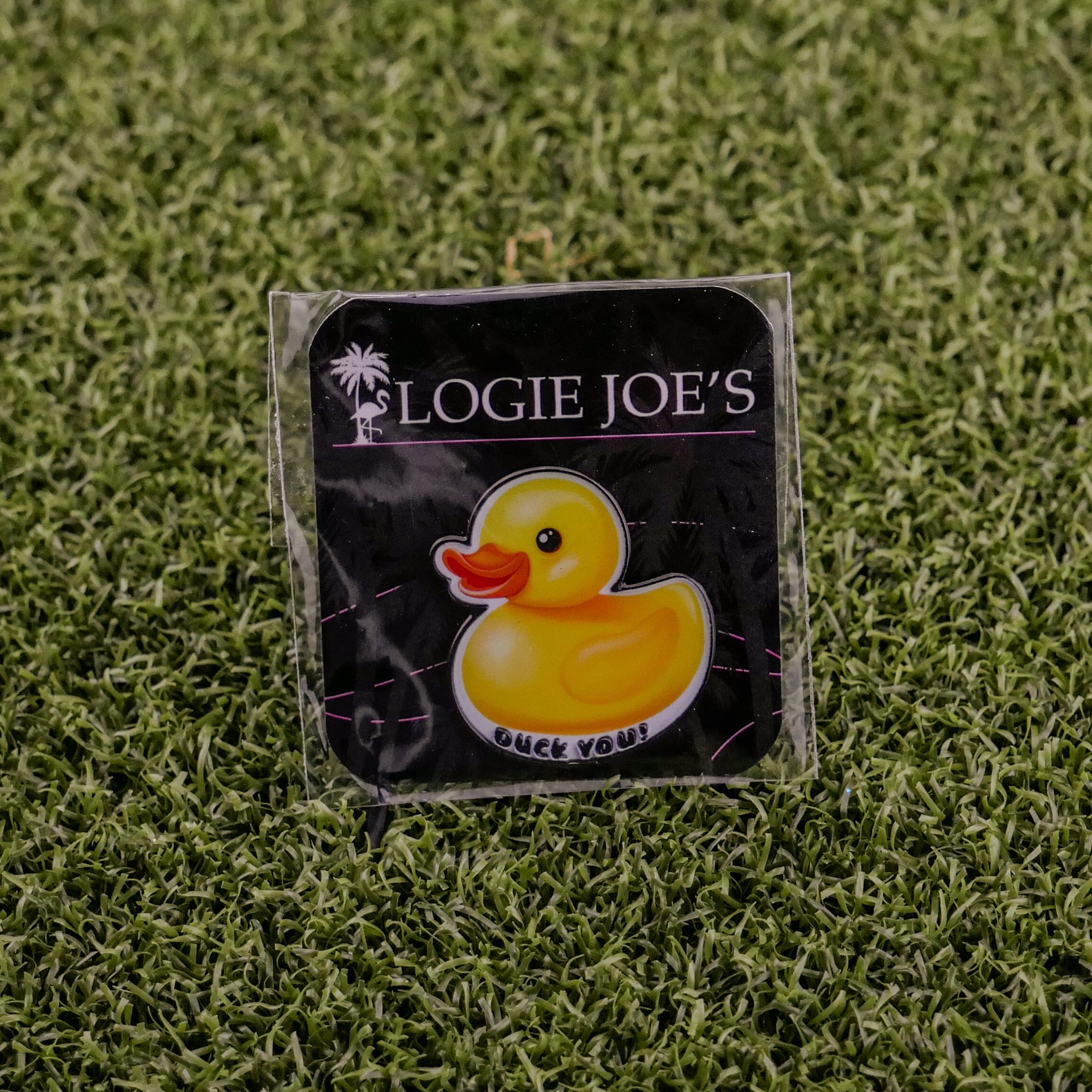 The Rubber Duck You pin from Logie Joe's Golf features a cartoon yellow duck and "QUACK YOU!" text, displayed on a black card—ideal for golf ball marker collectors. Set on green artificial grass for added style.