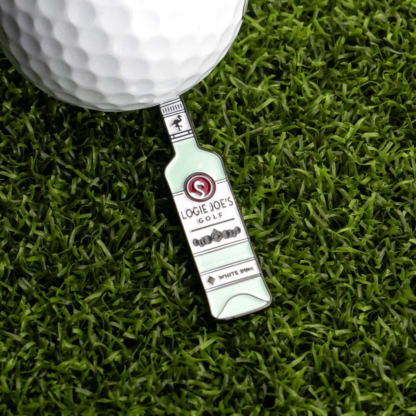 A golf ball rests on artificial grass next to a bottle-shaped metal White Rum Ball Marker from Logie Joe's Golf.