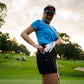 A female golfer in a blue shirt, black visor, and golf glove stands on the course, placing THE BYRD GOLF CLYP - Black Byrd by The Byrd, LLC in her shorts pocket; golf carts and trees are visible behind her.