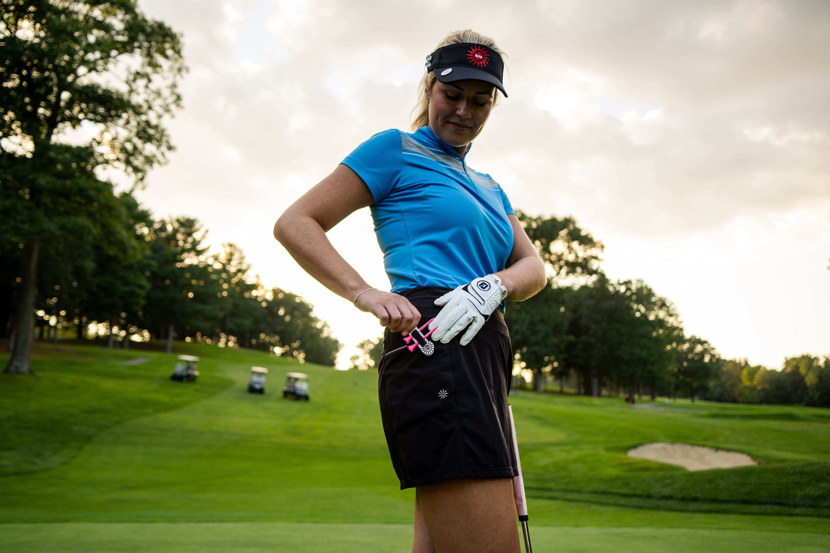 A female golfer in a blue shirt, black visor, and golf glove stands on the course, placing THE BYRD GOLF CLYP - Black Byrd by The Byrd, LLC in her shorts pocket; golf carts and trees are visible behind her.