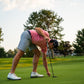 A golfer in a pink shirt and gray shorts bends to place a ball on the green, holding a putter. Featuring THE BYRD GOLF DIVOT CLYP by The Byrd, LLC—a golf multitool that organizes gear. Perfect for golf enthusiasts!.