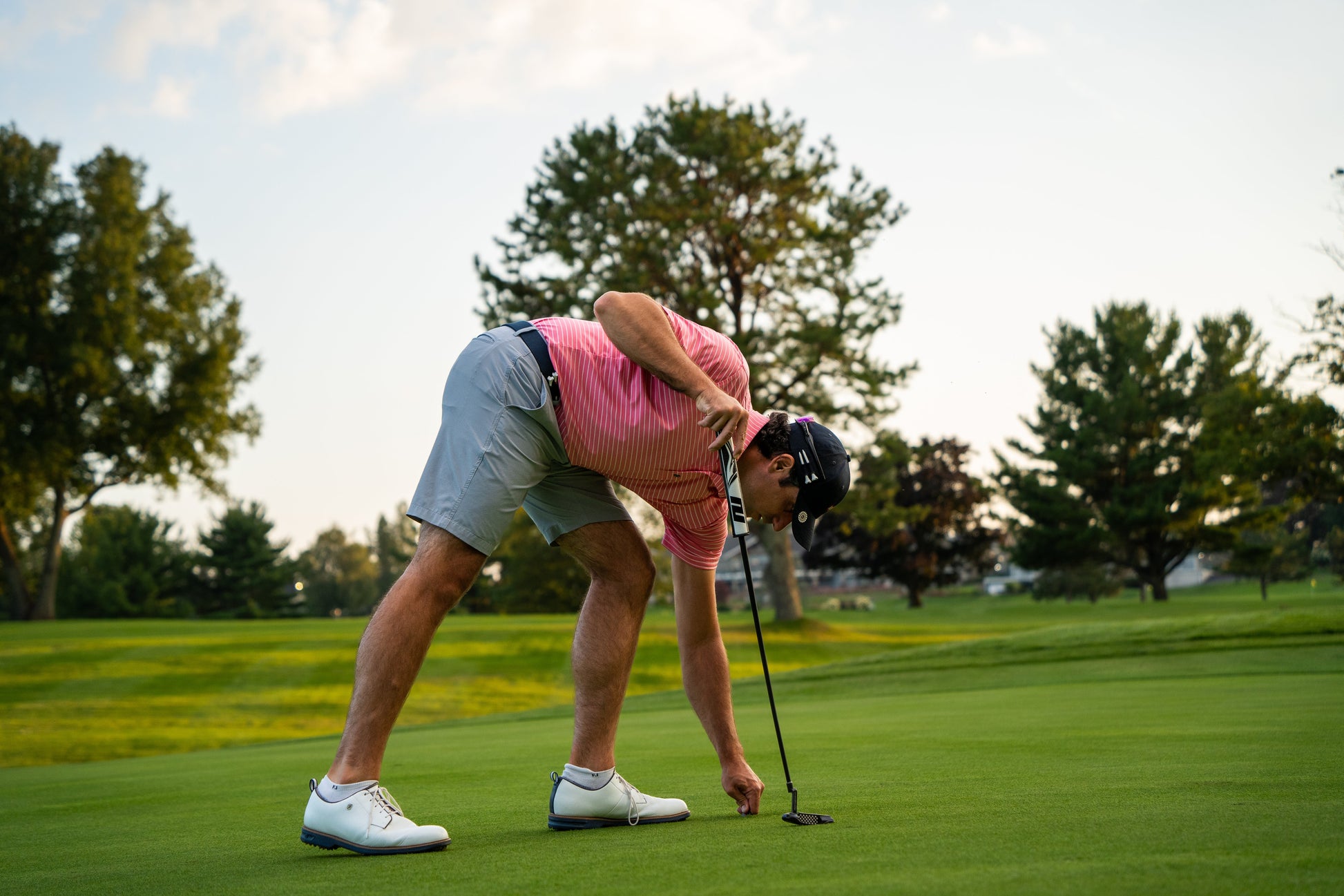 A golfer in a pink shirt and gray shorts bends to place a ball on the green, holding a putter. Featuring THE BYRD GOLF DIVOT CLYP by The Byrd, LLC—a golf multitool that organizes gear. Perfect for golf enthusiasts!.