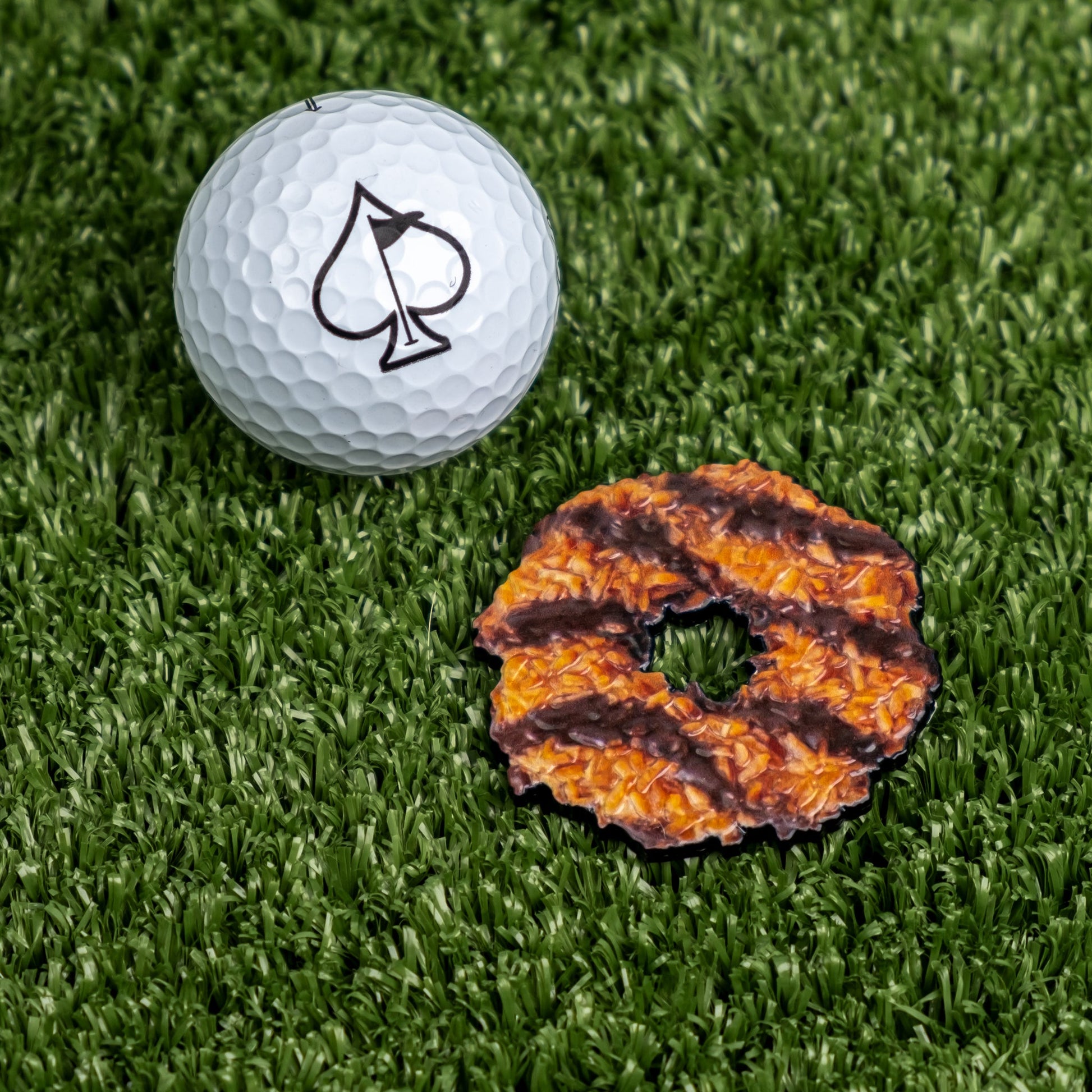 The Pins and Aces Coconut Fudge Cookie Ball Marker, featuring a black spade symbol, rests on artificial grass beside a Samoa-style caramel and coconut cookie.
