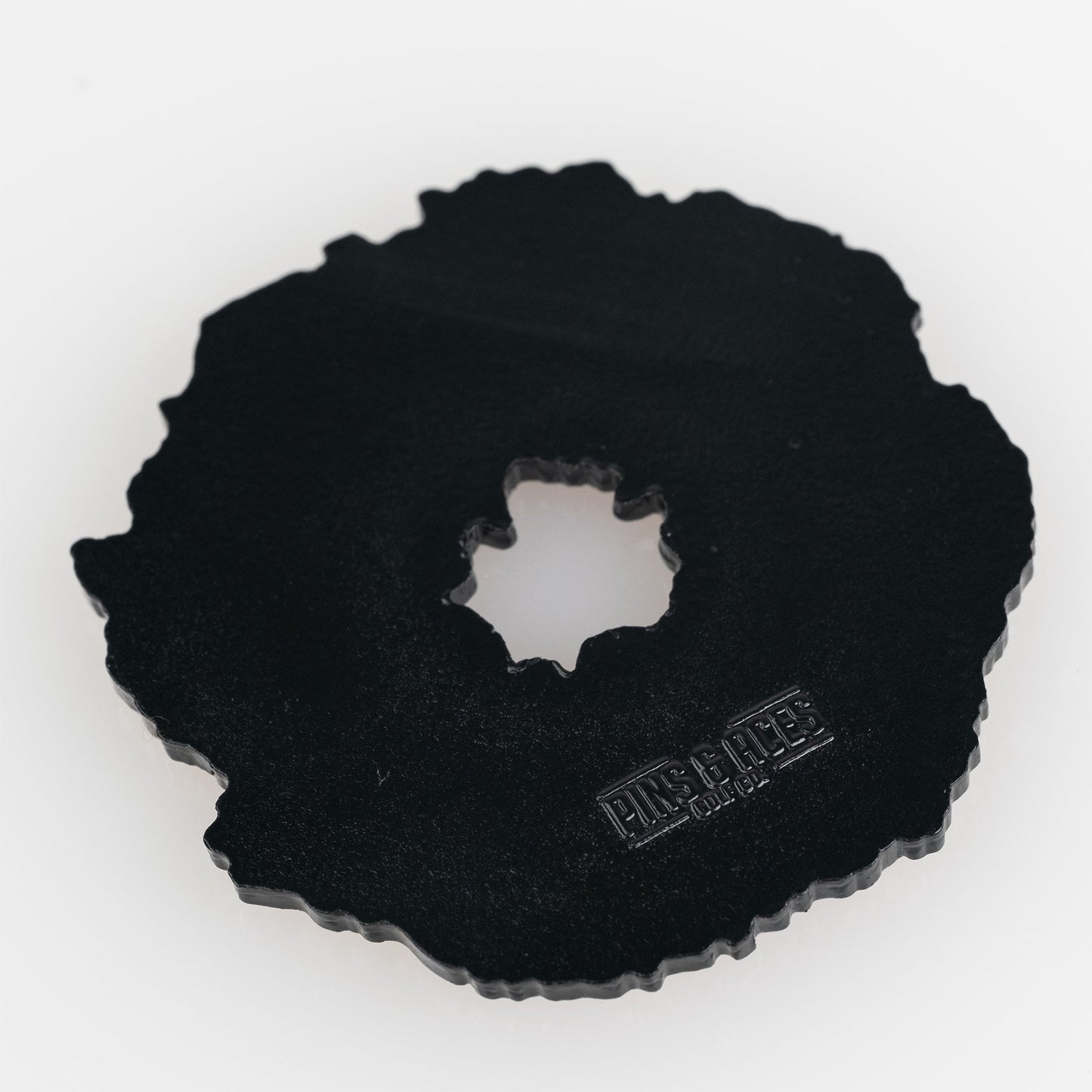 The Coconut Fudge Cookie Ball Marker by Pins and Aces is a black, irregular cast metal marker with a jagged edge, central hole, and matte finish. "PMS & RITE" plus other text are embossed on one side. Shown on a white background.