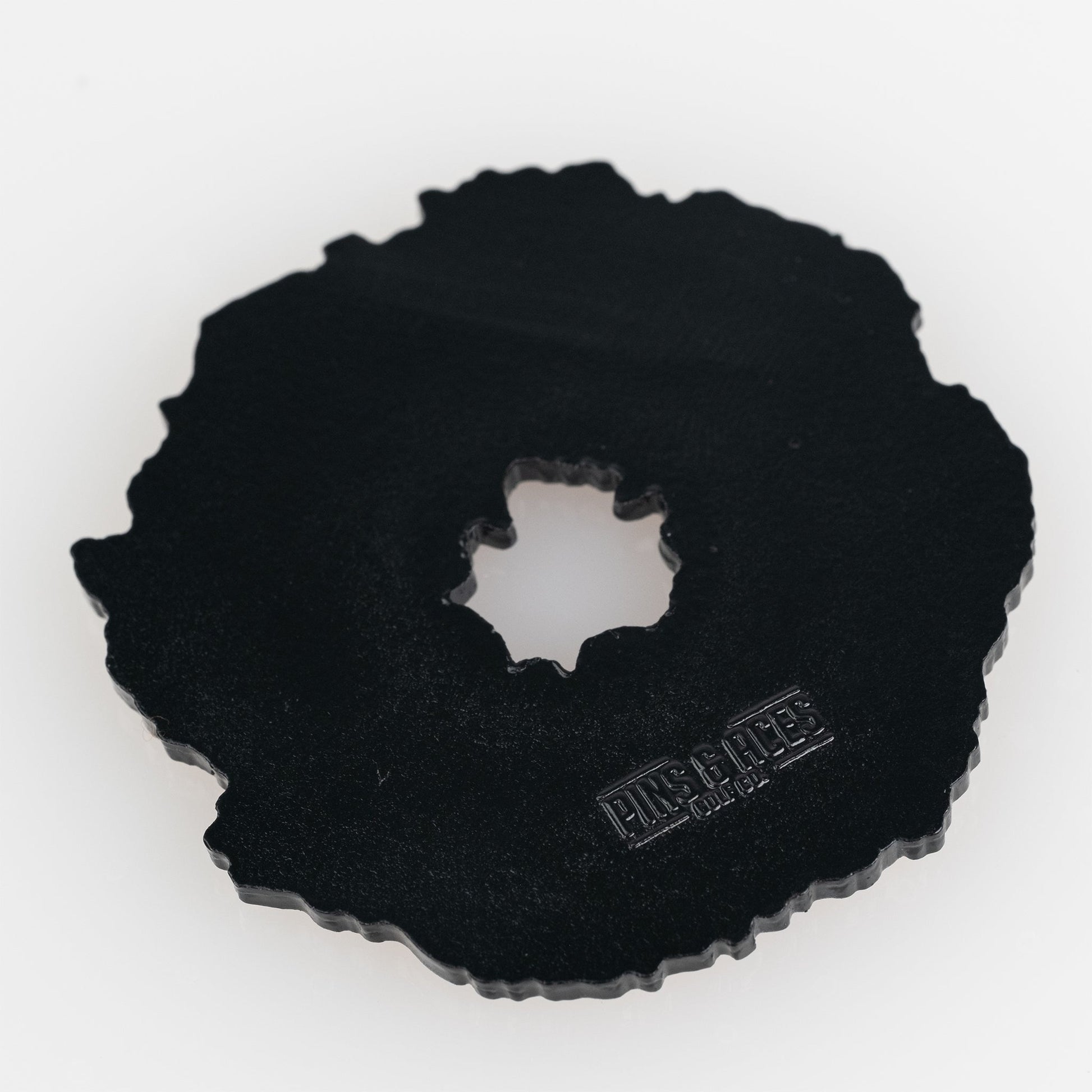 The Coconut Fudge Cookie Ball Marker by Pins and Aces is a black, irregular cast metal marker with a jagged edge, central hole, and matte finish. "PMS & RITE" plus other text are embossed on one side. Shown on a white background.