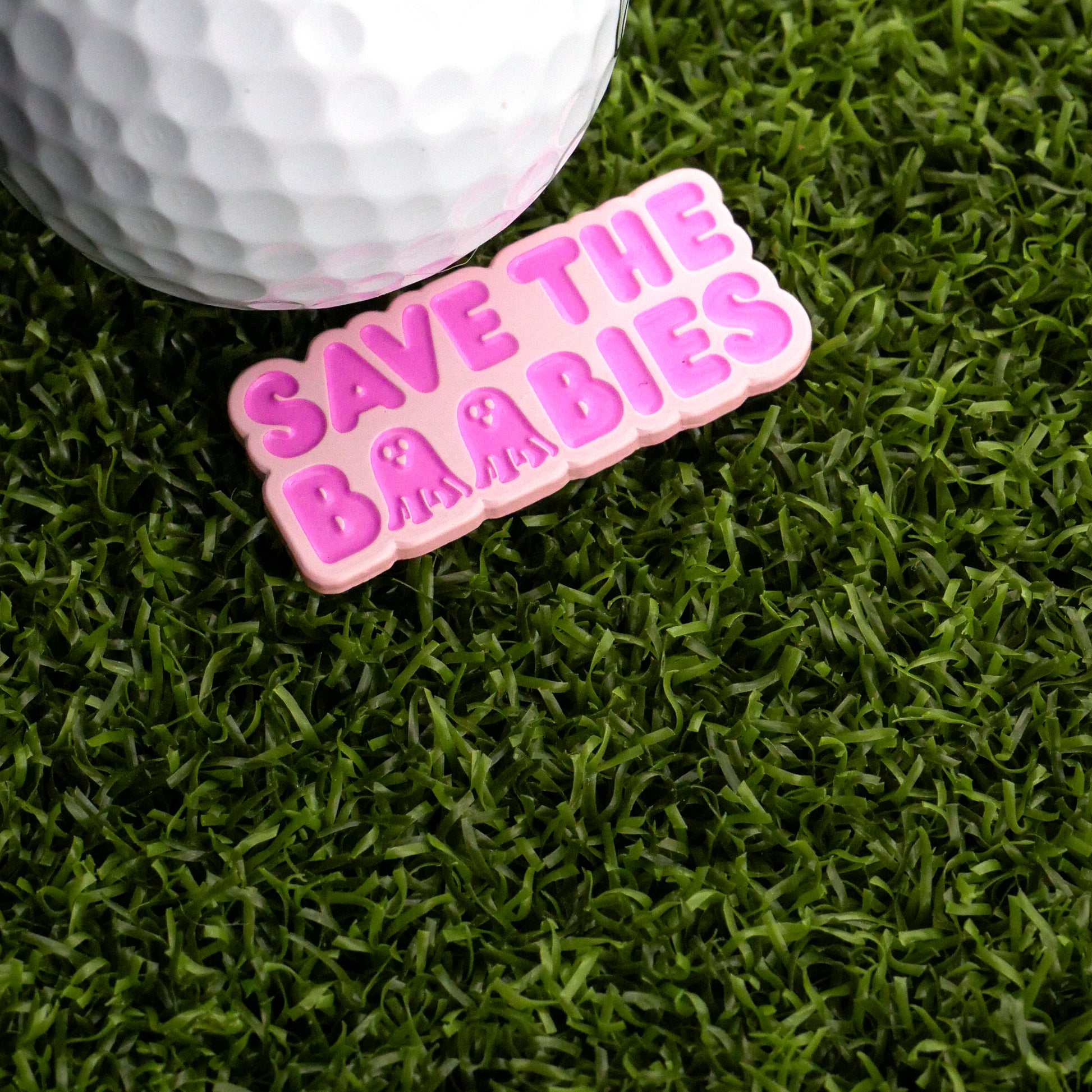 The Save The Boobies Ball Marker by Ballmarkers.com features a white golf ball and a pink octopus-inspired sign on green turf—an eye-catching awareness accessory for any putting green.