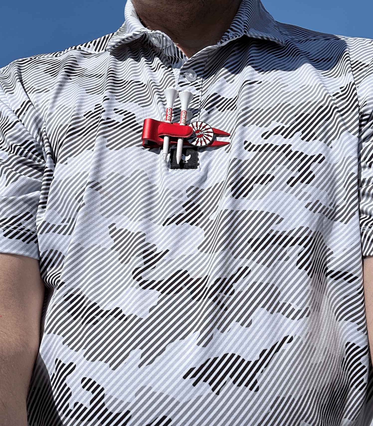 A person wears a patterned, short-sleeve shirt with THE BYRD GOLF DIVOT CLYP by The Byrd, LLC—an in-demand golf accessory with low stock—clipped to their chest, photographed from the neck down against a blue sky.