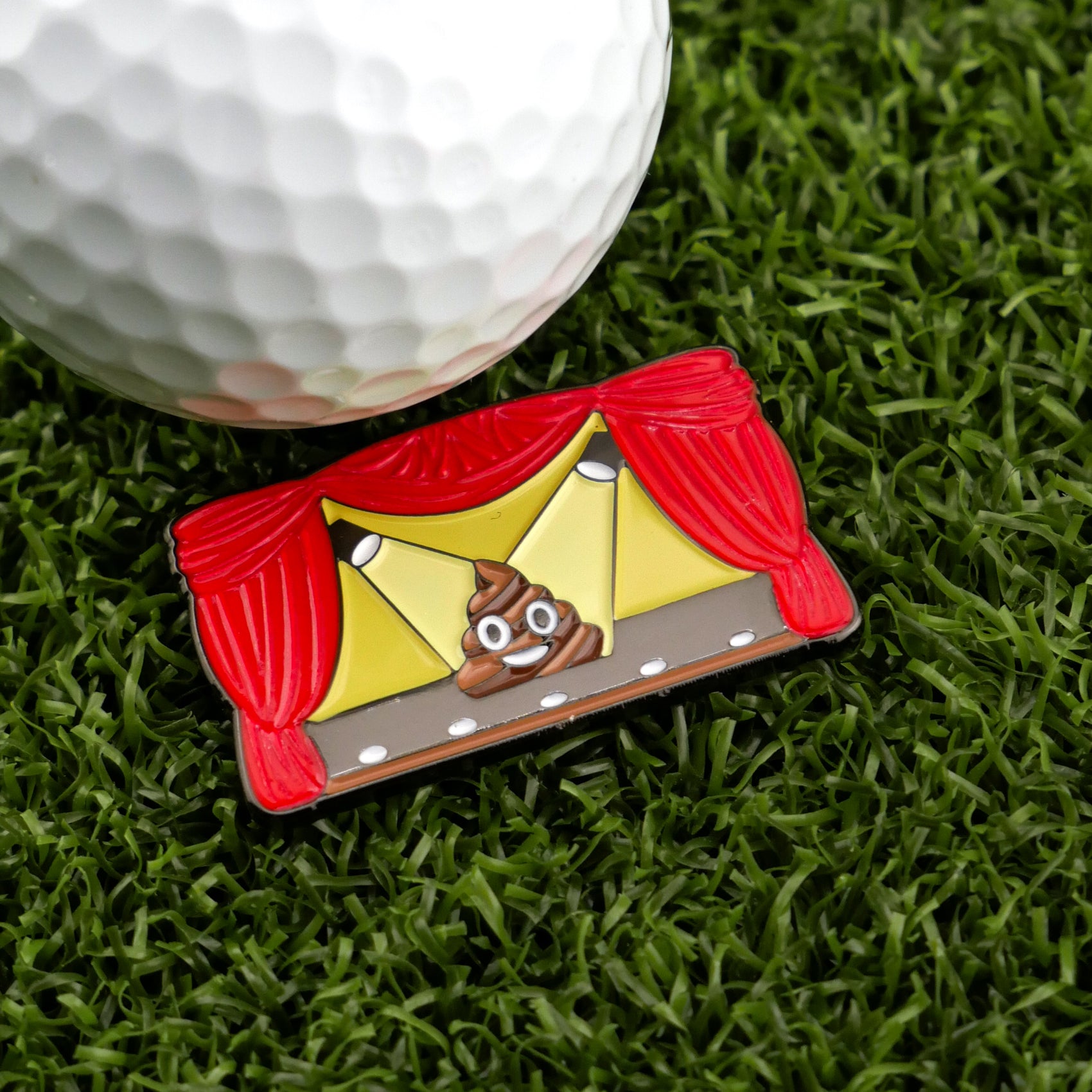 The Sh*t Show pin from Logie Joe's Golf features a poop emoji onstage with red curtains and yellow backdrop—perfect as a funny golf accessory or unique ball marker, shown next to a golf ball on artificial grass.