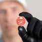 A person in black gloves holds Doubloon's The Silver Omen Antique, Color Edition—a personalized golf ball marker with a red coin, white skull and crossbones, and "SEIZE THE DAY"; their face is blurred in the background.