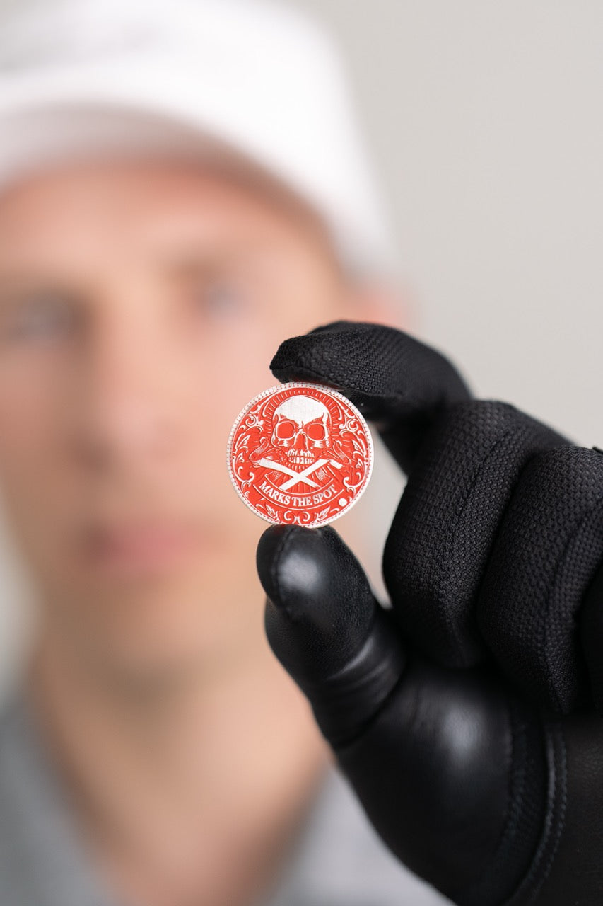 A person in black gloves holds Doubloon's The Silver Omen Antique, Color Edition—a personalized golf ball marker with a red coin, white skull and crossbones, and "SEIZE THE DAY"; their face is blurred in the background.