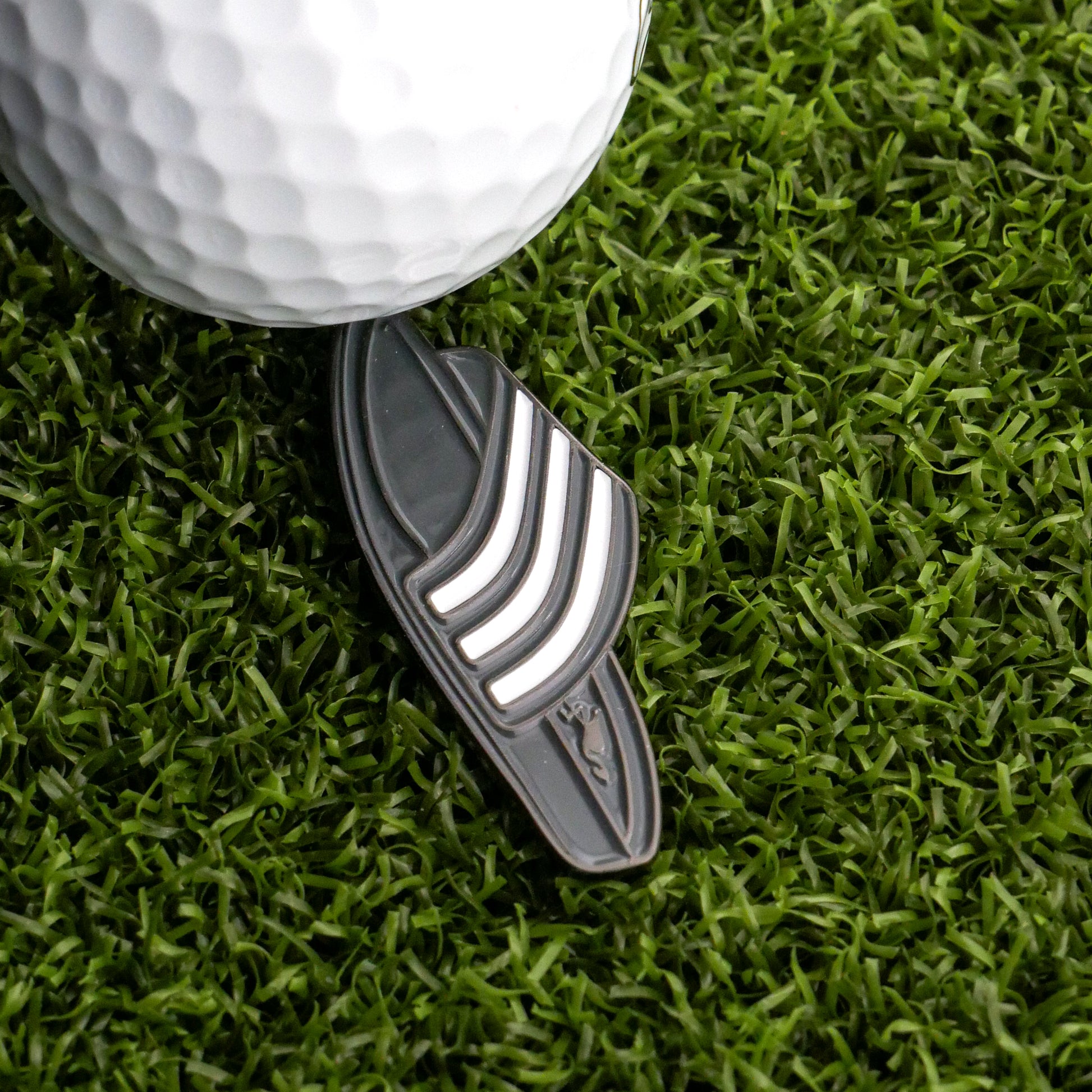 A close-up of a white golf ball on green grass next to the Ballmarkers.com Slide Sandal Ball Marker, a black marker with three white stripes, ready for the putting game.