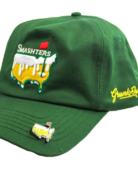 The Grunk Dolfer Smashters Ball Marker Ball Marker is a green cap with a golf course-and-beer patch labeled SMASHTERS, yellow side script, and a pin on the bill that serves as a unique golf ball marker—an ideal golf gift.
