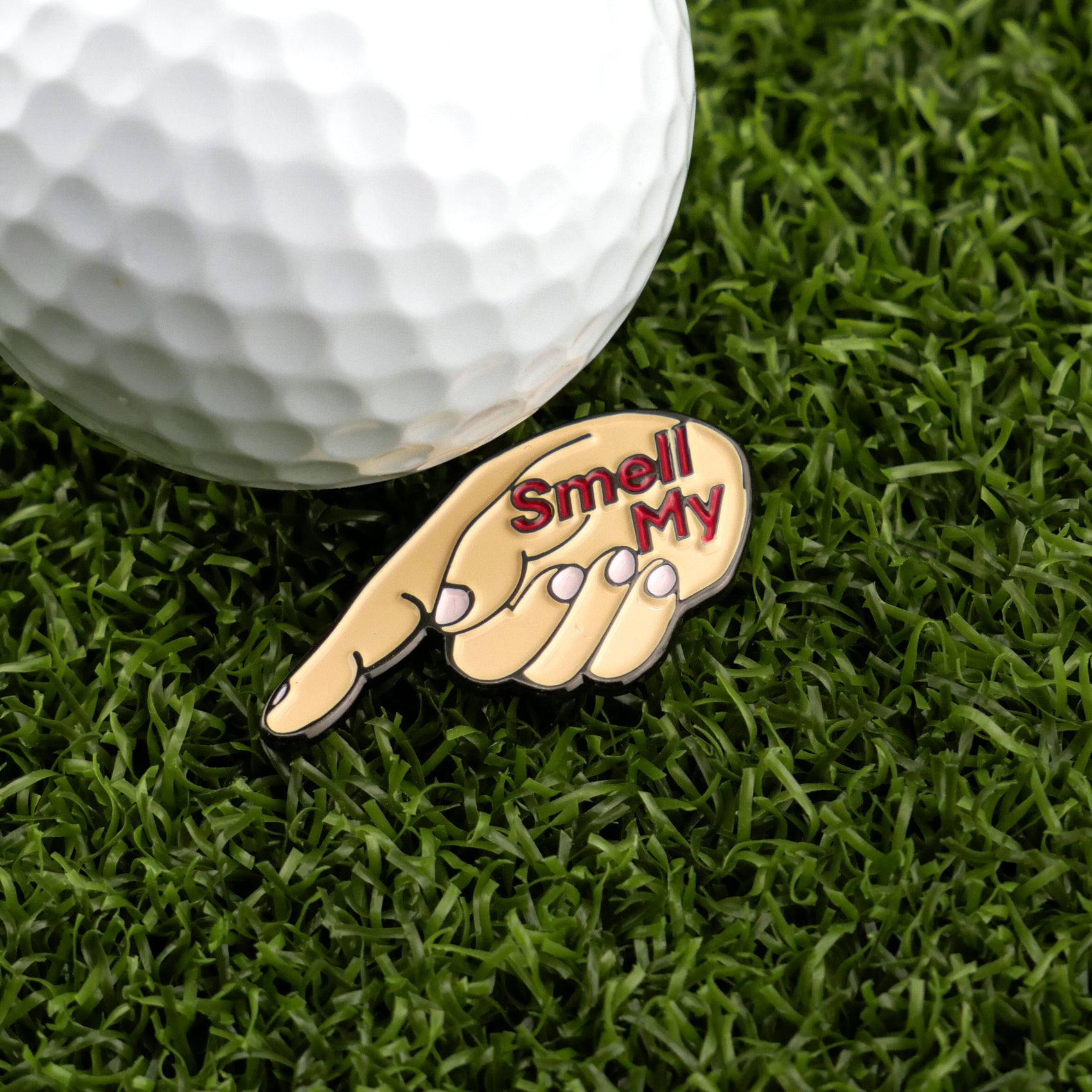 A golf ball rests on green grass beside Logie Joe's Golf’s Smell My Finger ball marker—a hand-shaped marker with "Smell My" in red.