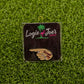The Smell My Finger ball marker by Logie Joe’s Golf, shaped like a pointing hand with “Smell My,” rests on green turf and comes displayed on a branded card—a hilarious upgrade to your premium golf accessories.