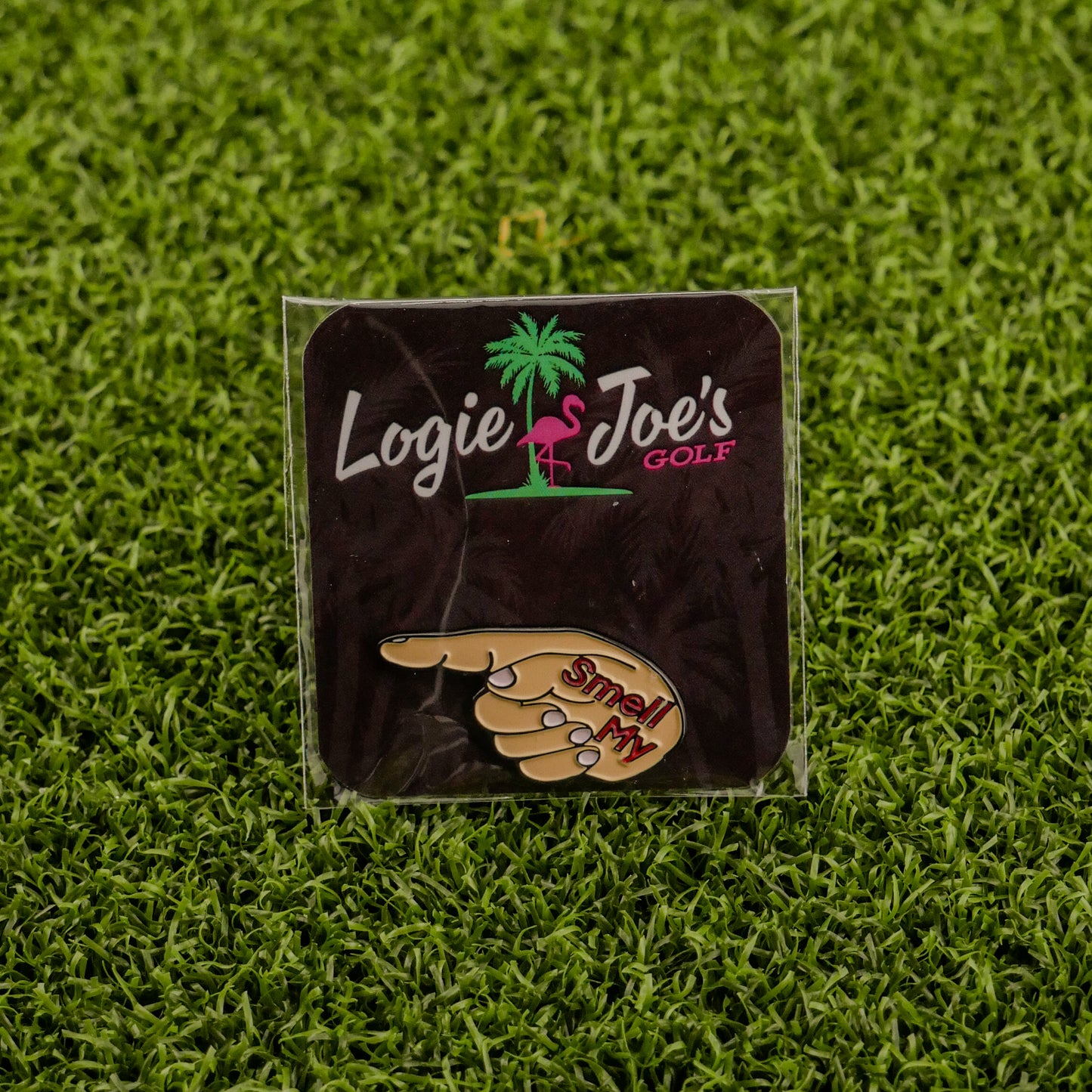 The Smell My Finger ball marker by Logie Joe’s Golf, shaped like a pointing hand with “Smell My,” rests on green turf and comes displayed on a branded card—a hilarious upgrade to your premium golf accessories.