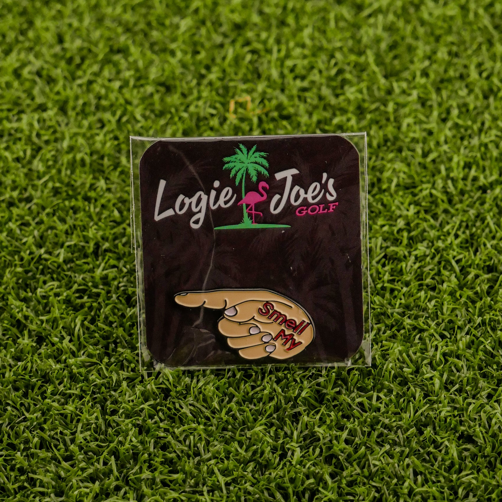 The Smell My Finger ball marker by Logie Joe’s Golf, shaped like a pointing hand with “Smell My,” rests on green turf and comes displayed on a branded card—a hilarious upgrade to your premium golf accessories.