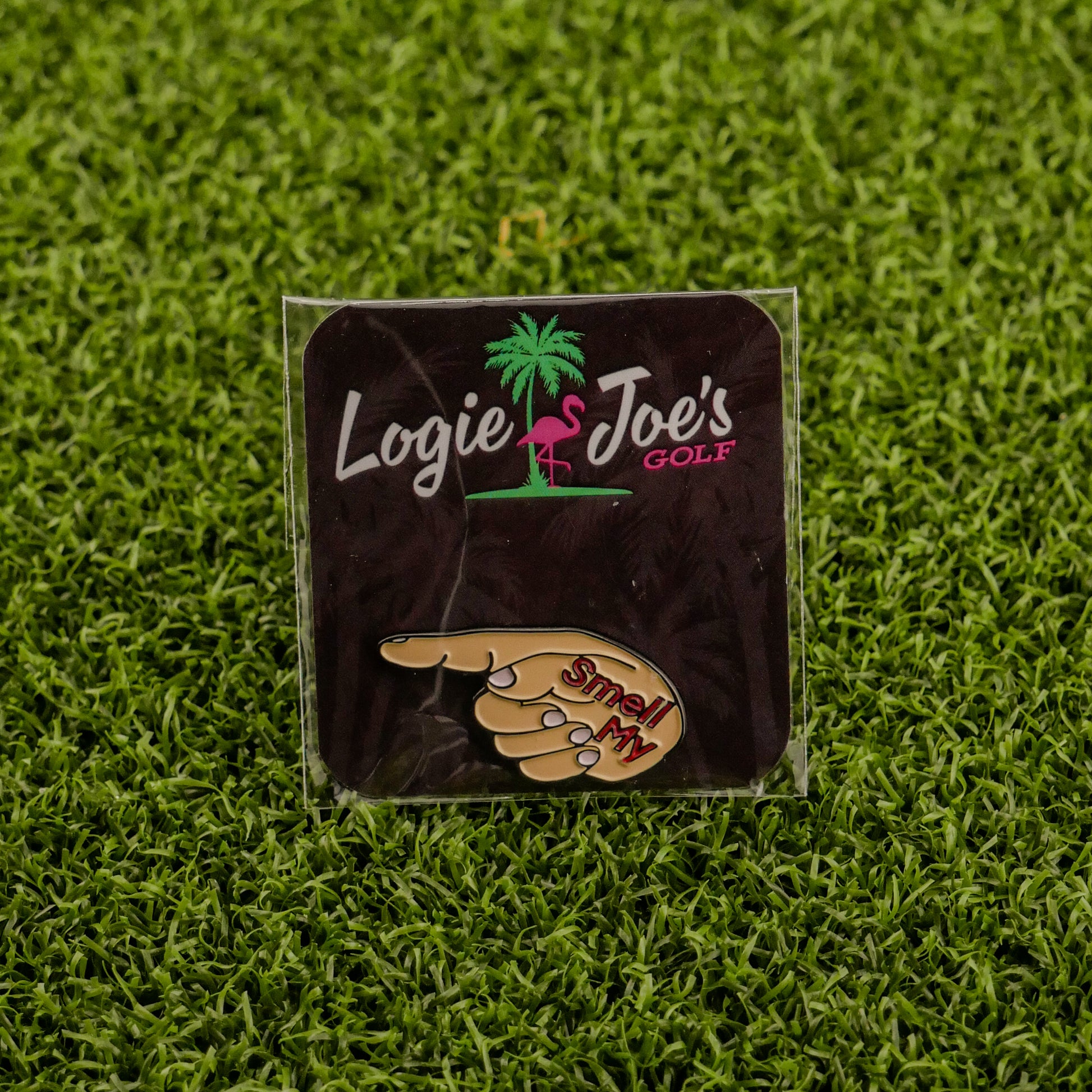 The Smell My Finger ball marker by Logie Joe’s Golf, shaped like a pointing hand with “Smell My,” rests on green turf and comes displayed on a branded card—a hilarious upgrade to your premium golf accessories.