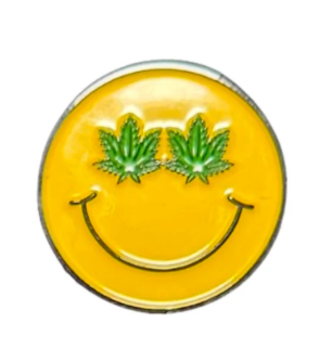 The Smiley Stoned Ball Marker from Golf Gear Heaven features a yellow smiley face with green cannabis leaf eyes—ideal for golf lovers to use on the course or accessorize your Tee Bumps gear.