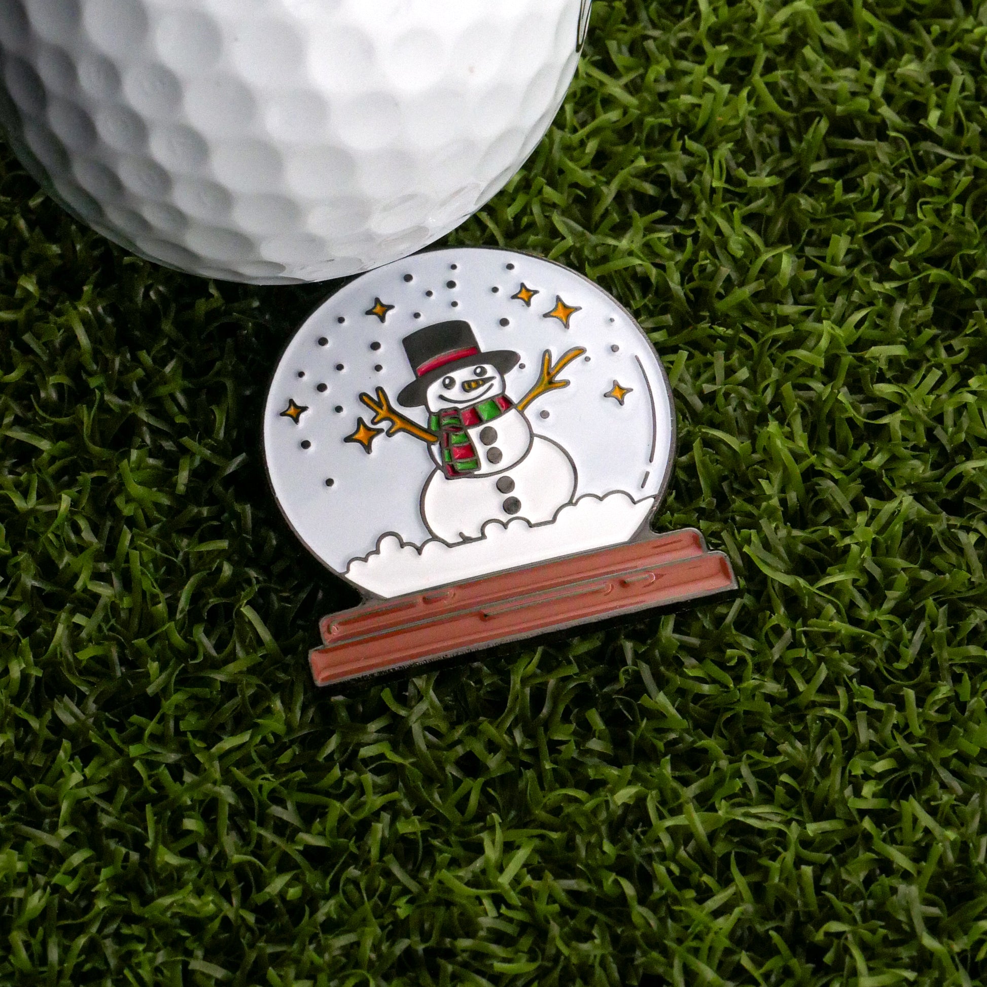 The Frosty the Snow Globe Ball Marker by Ballmarkers.com features a smiling snowman in a black hat and green scarf, sitting on artificial grass next to a white golf ball—perfect for adding festive cheer to your game.
