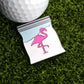 A Snowcane Baggie Ball Marker by Logie Joe's Golf—a novelty enamel pin shaped like a plastic bag with a pink flamingo—sits next to a golf ball on artificial grass, creating a unique golf accessory.