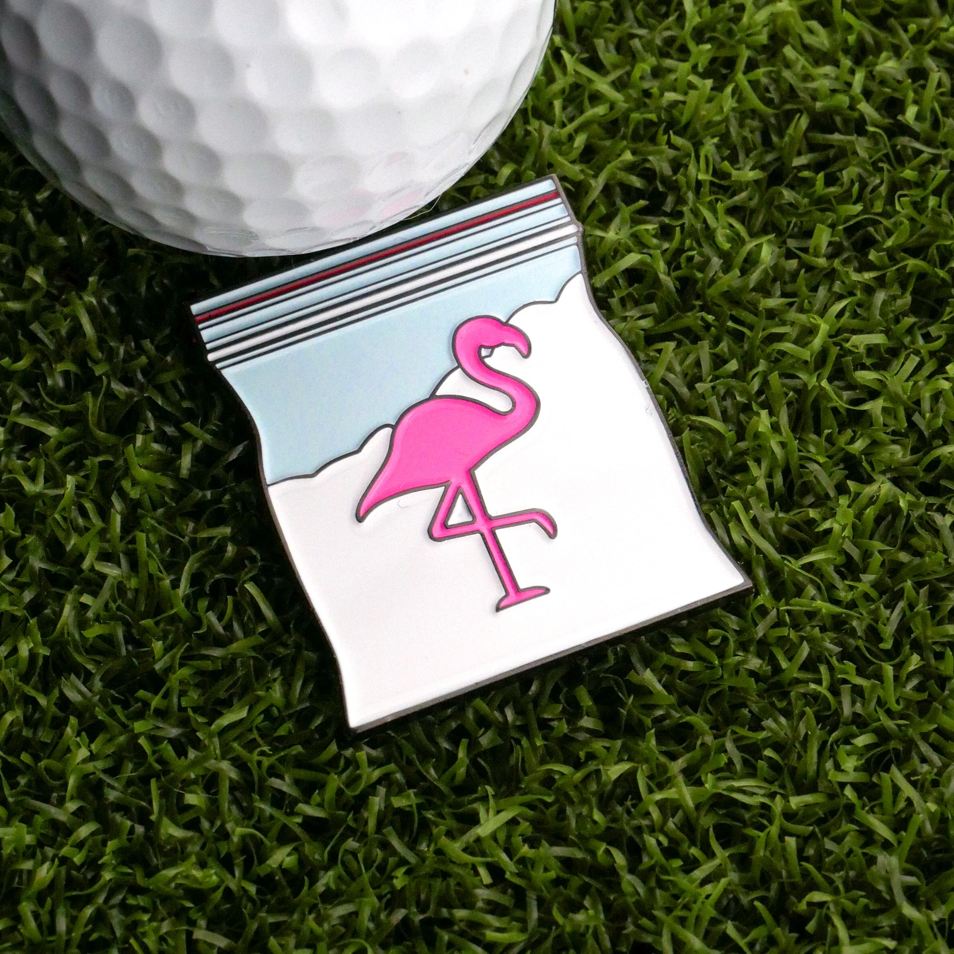 A Snowcane Baggie Ball Marker by Logie Joe's Golf—a novelty enamel pin shaped like a plastic bag with a pink flamingo—sits next to a golf ball on artificial grass, creating a unique golf accessory.