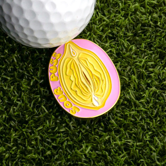 The Sour Puss Ball Marker by Logie Joe's Golf is a pink and yellow lemon-inspired oval pin with "Sour Puss" and a stylized vulva, shown on artificial grass beside a golf ball for a cheeky touch.