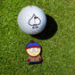 A white golf ball with a black spade symbol sits on green grass next to the South Park Ball Marker - Stan, featuring Stan in a blue and red hat and brown coat, from the Pins and Aces collection.