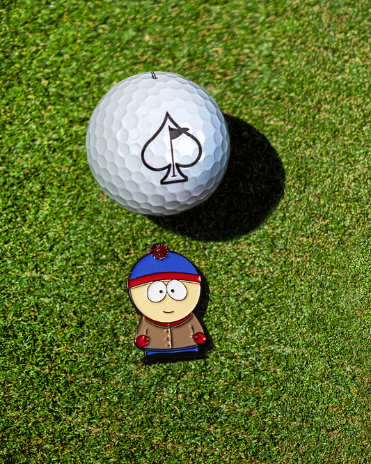 A white golf ball with a black spade symbol sits on green grass next to the South Park Ball Marker - Stan, featuring Stan in a blue and red hat and brown coat, from the Pins and Aces collection.