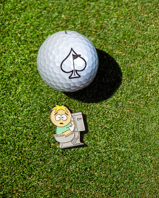 A golf ball with a spade symbol sits on the grass beside the Pins and Aces South Park Ball Marker - Butters, featuring a worried child on a toilet.