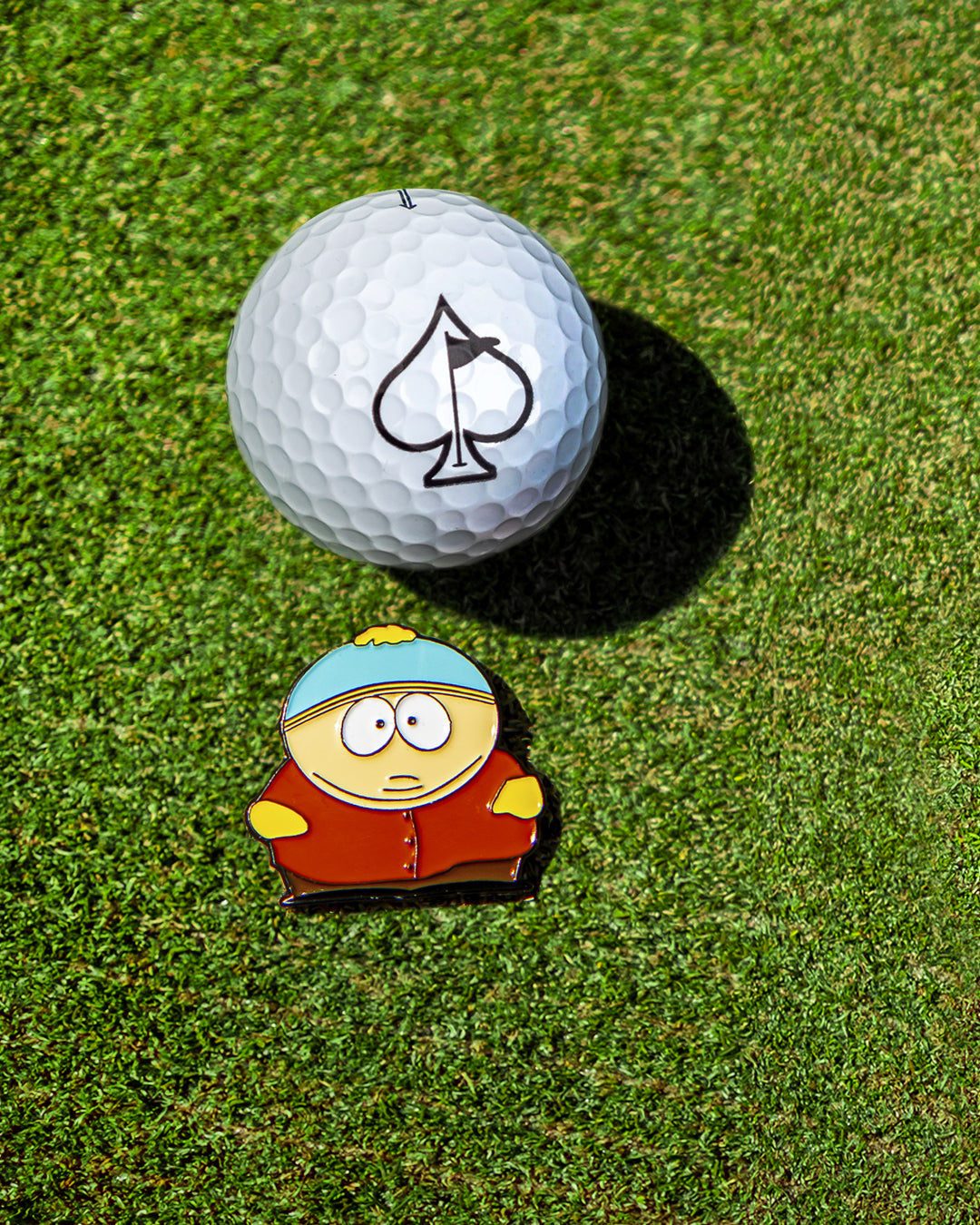 The Pins and Aces South Park Ball Marker - Cartman features a cartoon Cartman next to a spade symbol on green grass, ideal for South Park golf fans or collectors of premium Pins and Aces ball markers.