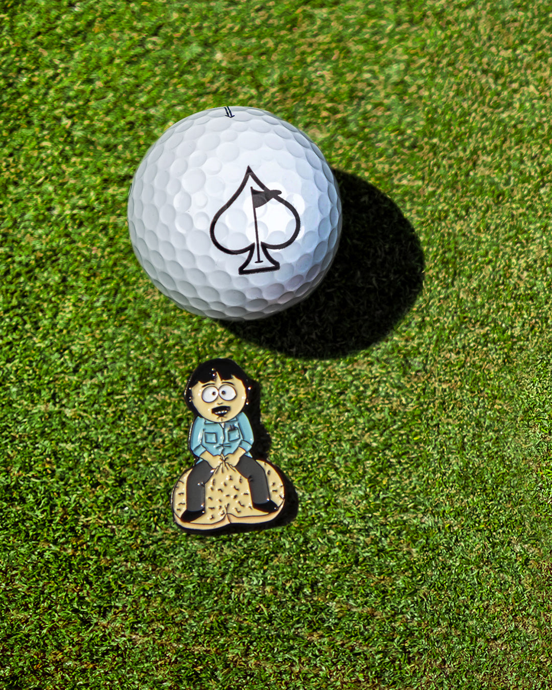 The South Park Ball Marker - Randy by Pins and Aces features a surprised cartoon character on sand and a spade on a white golf ball—fun, unique additions to your premium ball markers lineup.
