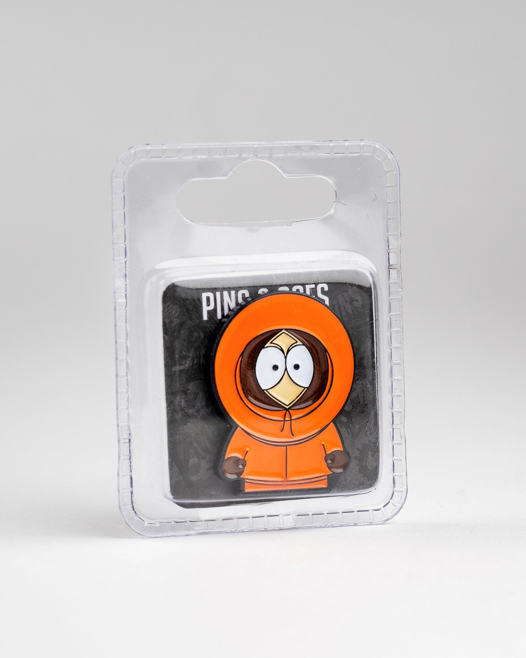 The Pins and Aces South Park Ball Marker - Kenny is displayed in orange hoodie packaging with large eyes and a covered face, on a black backing card in a clear plastic case against a plain white background.