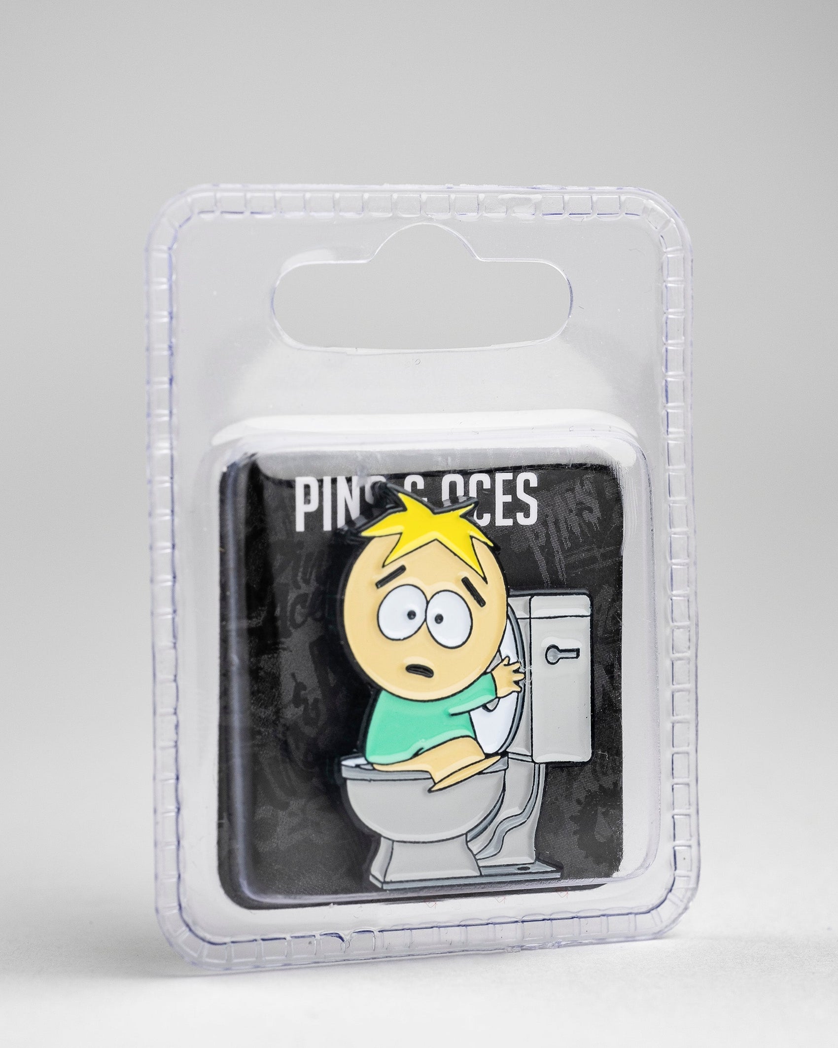 The South Park Ball Marker - Butters by Pins and Aces features a blond character, in South Park golf gear style, surprised on a toilet and comes in plastic packaging with a gray background.