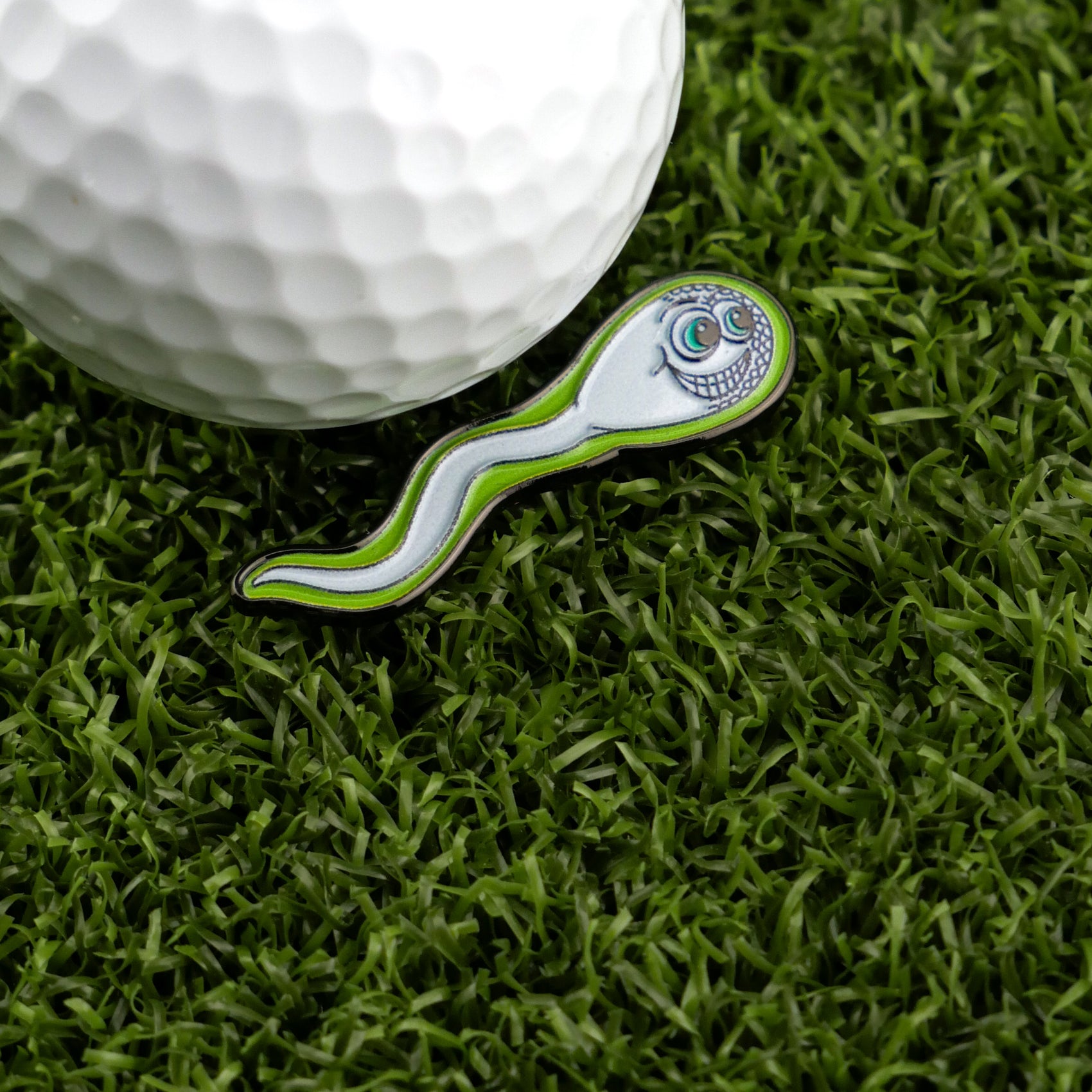 The Logie Joe's Golf Sperm Cell pin features a smiling face and cartoonish design, sitting on green turf beside a golf ball—making it a fun and unique addition to your premium golf accessories as a memorable ball marker.