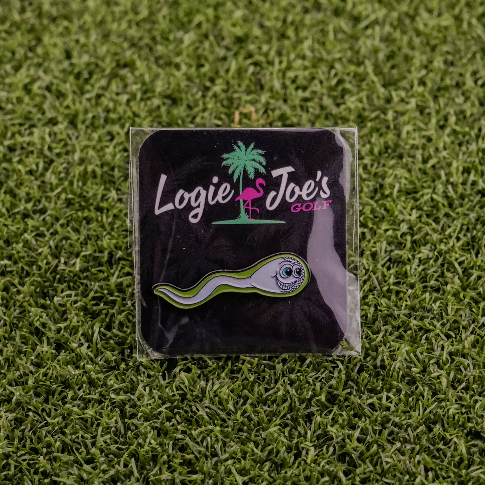 The Sperm Cell pin by Logie Joe's Golf is a playful golf ball marker featuring a cartoon sperm design, displayed on a branded card with a pink flamingo, and set on artificial green grass—ideal for premium golf accessory fans.