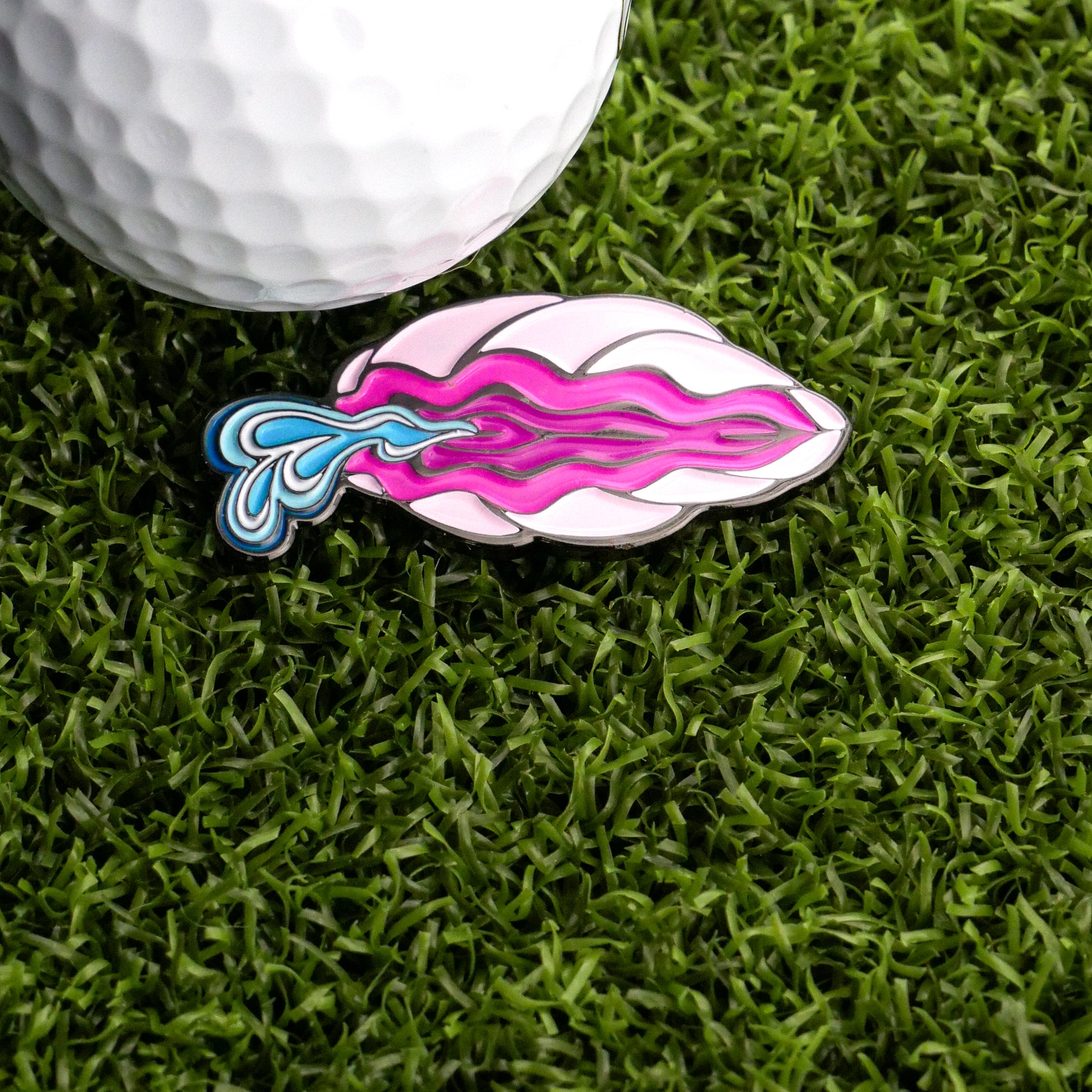 A golf ball rests on green artificial grass beside the Squirting Clam Ball Marker by Bunkers & Rough—a vibrant enamel pin shaped like lips with pink, purple waves, and a blue splash that’s sure to spark conversation on the course.