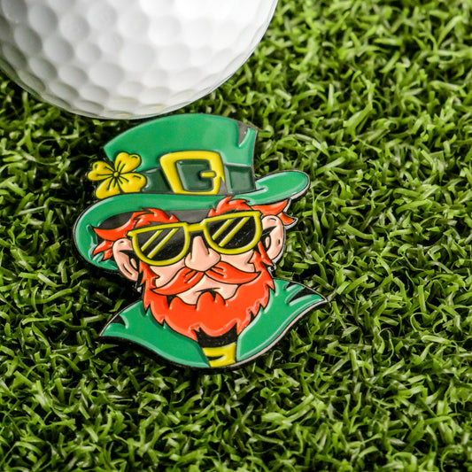 The Leprechaun Ball Marker by Ballmarkers.com features a red-haired cartoon leprechaun in green clothes and sunglasses, relaxing on grass near a golf ball—a playful St. Patrick's Day design perfect for any golfer.