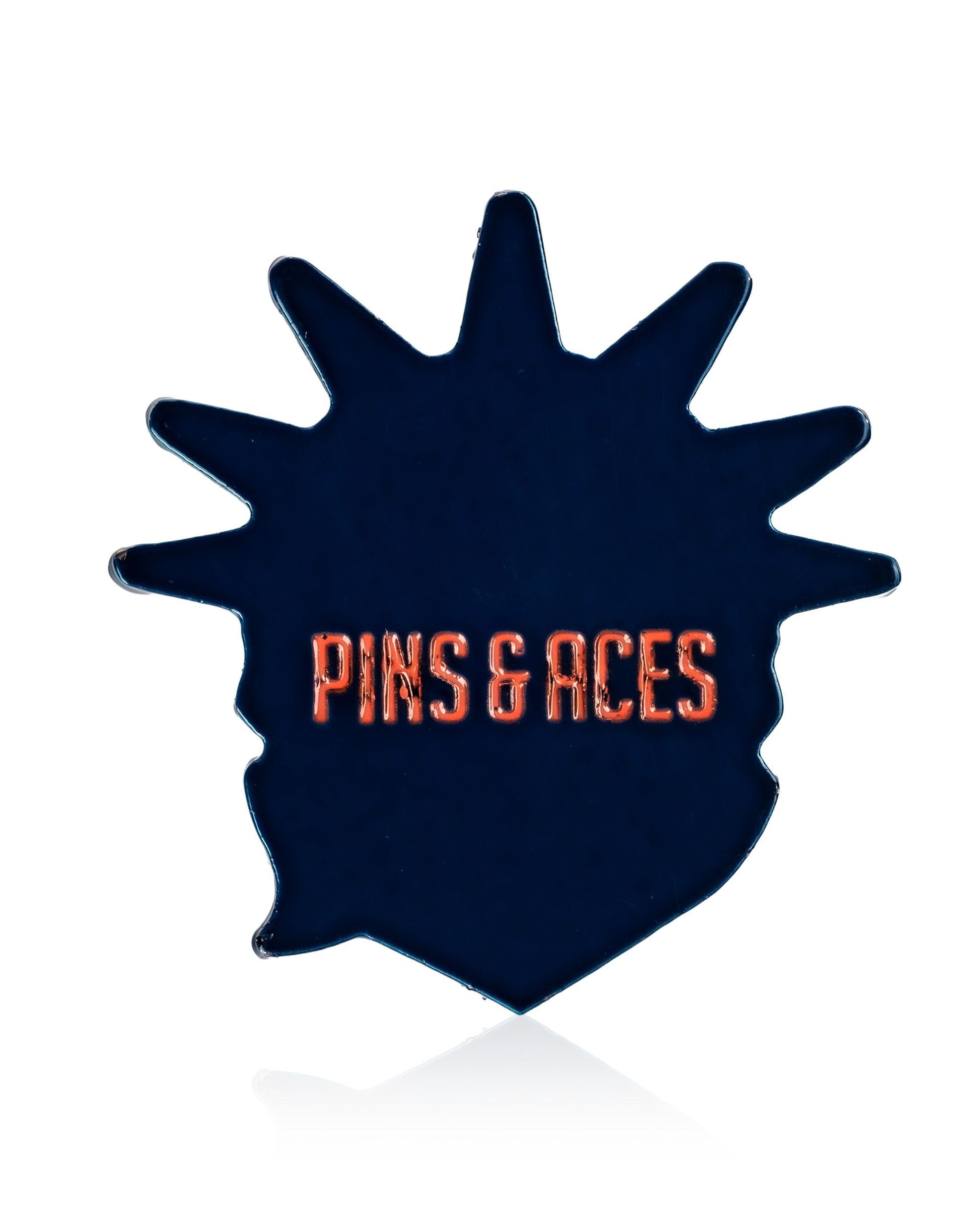 A dark, irregularly shaped stainless steel golf marker by Pins and Aces features “Statue of Liberty” in bold orange at the center. The background is plain white.