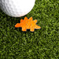 A white golf ball rests on artificial green grass beside a Ballmarkers.com Stegosaurus Dinosaur Nugget Ball Marker shaped like an orange stegosaurus.
