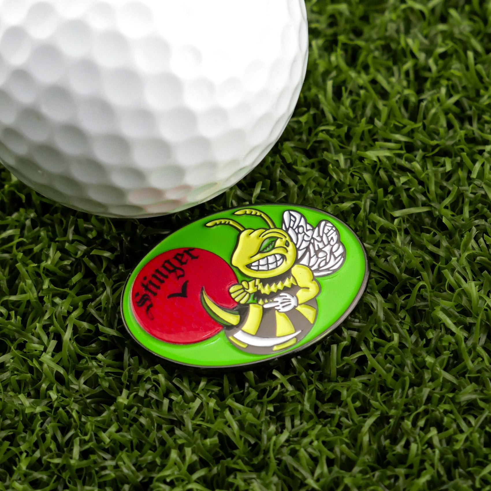 A close-up of a golf ball on artificial grass next to Logie Joe’s Golf "Stinger" marker, featuring a grinning yellow bee with a tee, the word “Stinger,” and a red ball—ideal for fans of premium golf accessories.