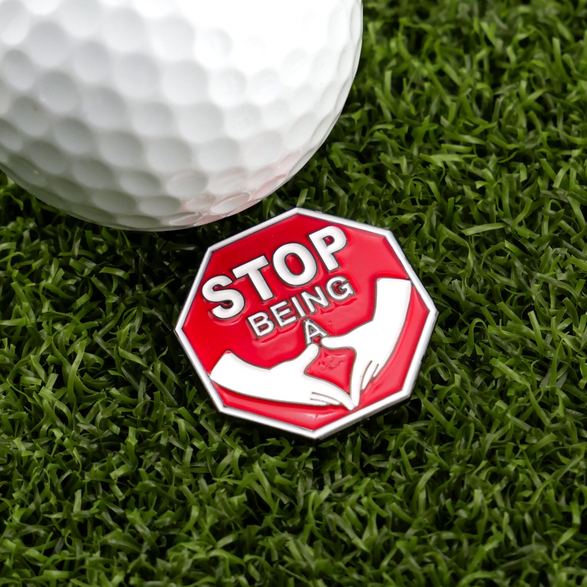 Logie Joe's Golf "Stop Being a P*%#!" ball marker—a red, octagonal accessory with cartoon hands—adds humor and style to your premium golf gear collection. Shown on green grass beside a white golf ball.