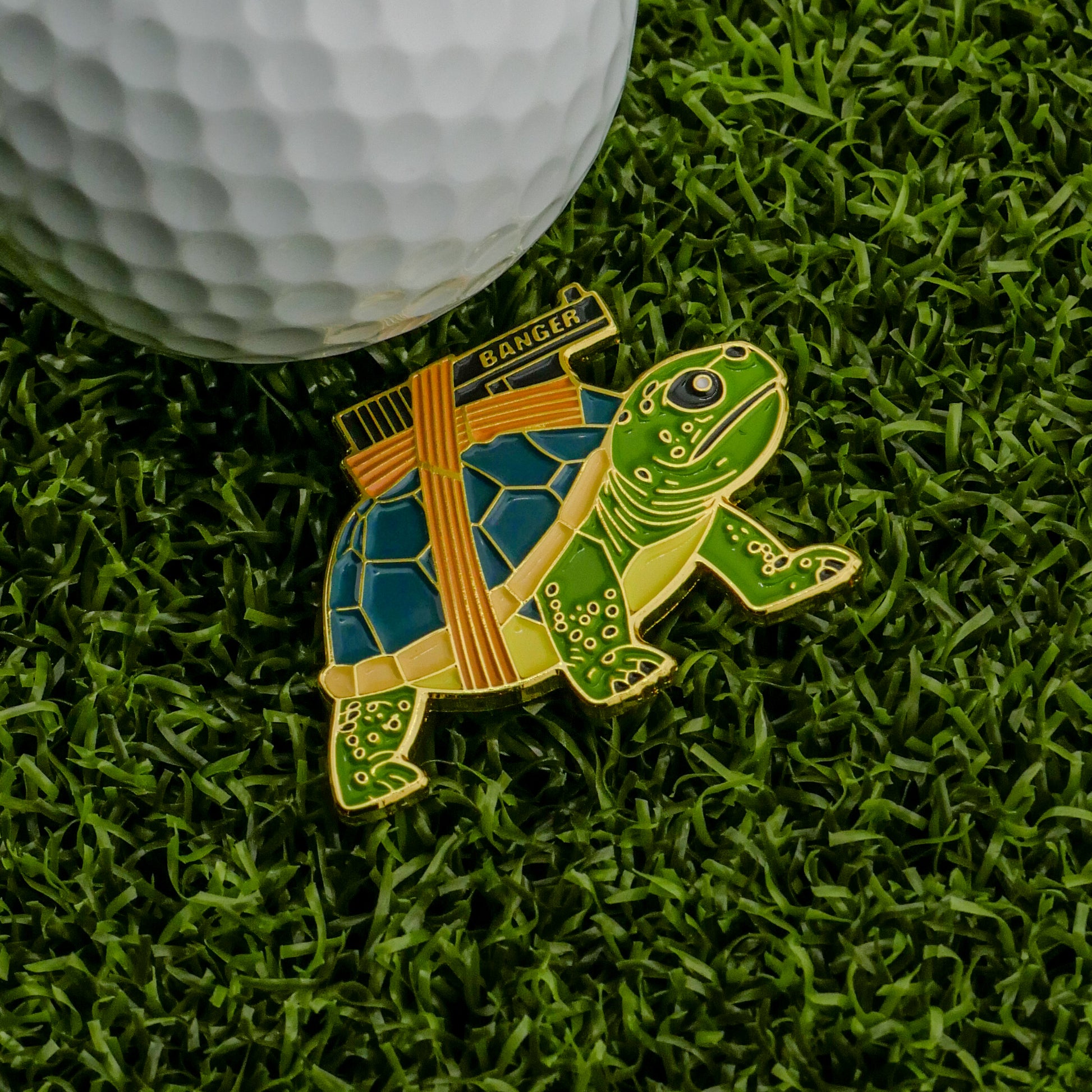 The Strapped Turtle Ball Marker from Logie Joe's Golf, featuring a blue and green shell labeled BANGER, sits on green artificial grass beside a white golf ball.