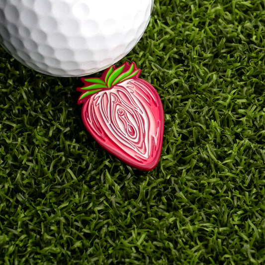 A close-up of a golf ball on green artificial grass next to Logie Joe's Golf Strawberry Vagina Ball Marker, featuring a detailed and stylized design.