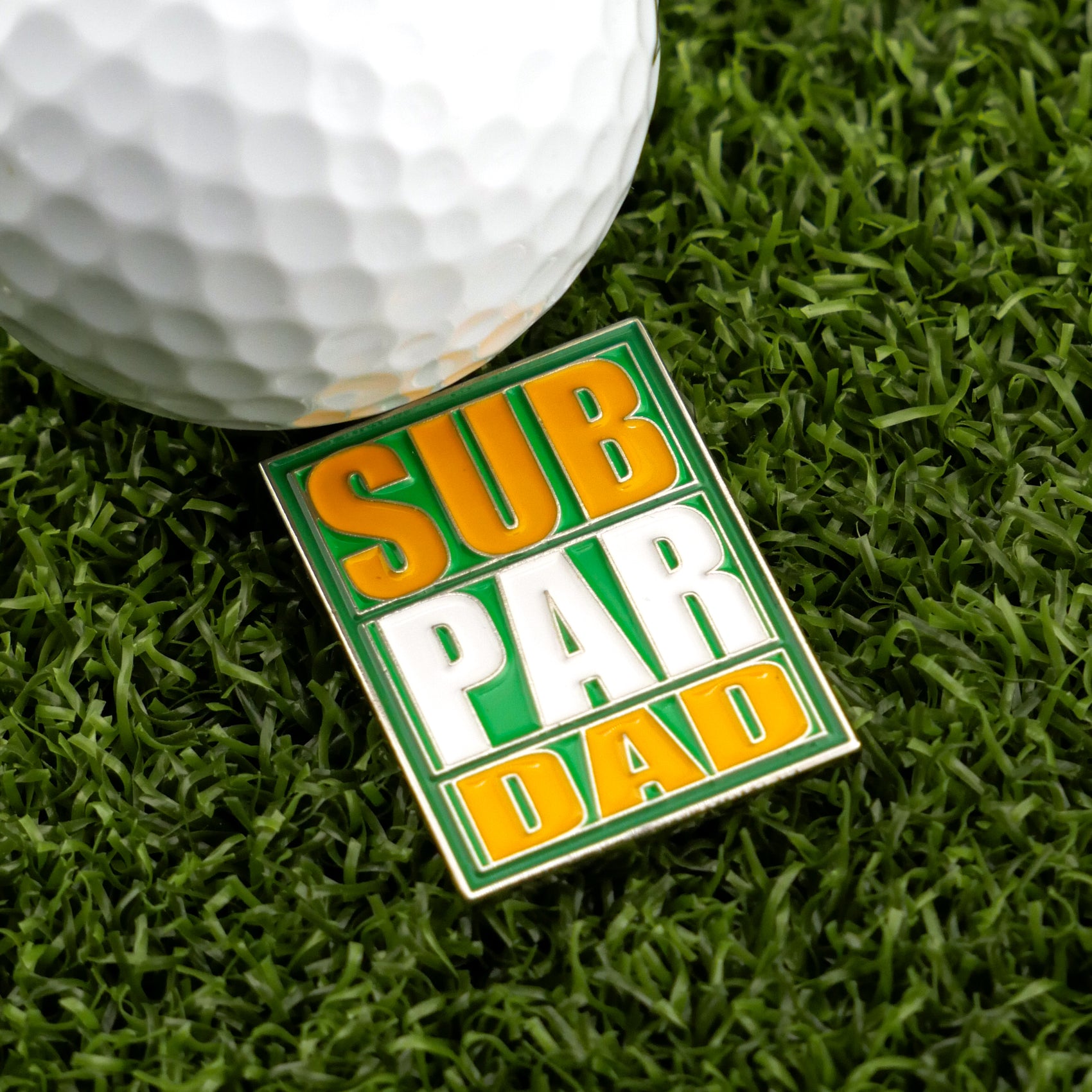 The Logie Joe's Golf "Sub Par Dad" rectangular pin with bold green, yellow, and white text rests on artificial grass beside a golf ball—an ideal personalized accessory or unique ball marker for any golf-loving dad.