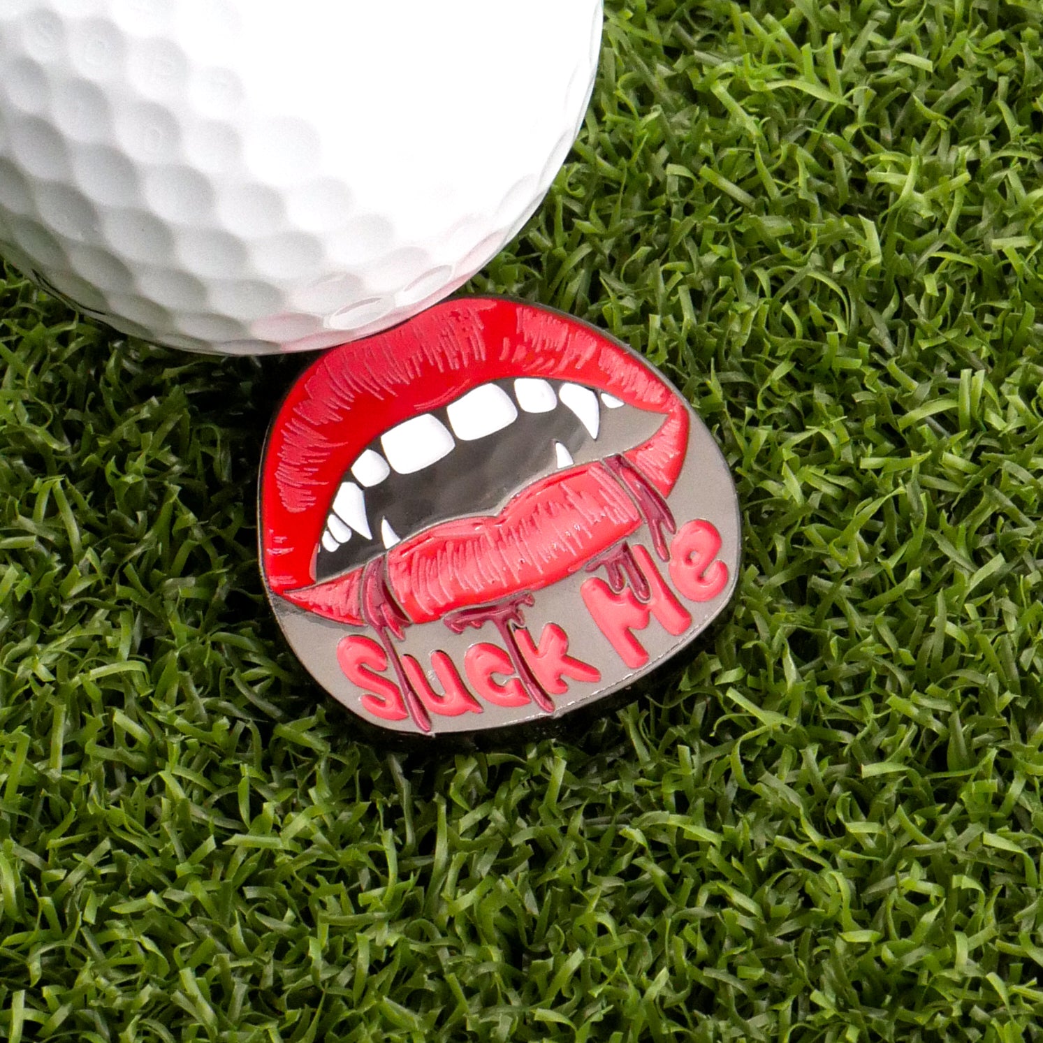 A golf ball on artificial grass sits beside Logie Joe's Golf Suck Me Ball Marker, a metal marker shaped like red lips with fangs and the phrase "Suck Me" in pink letters.